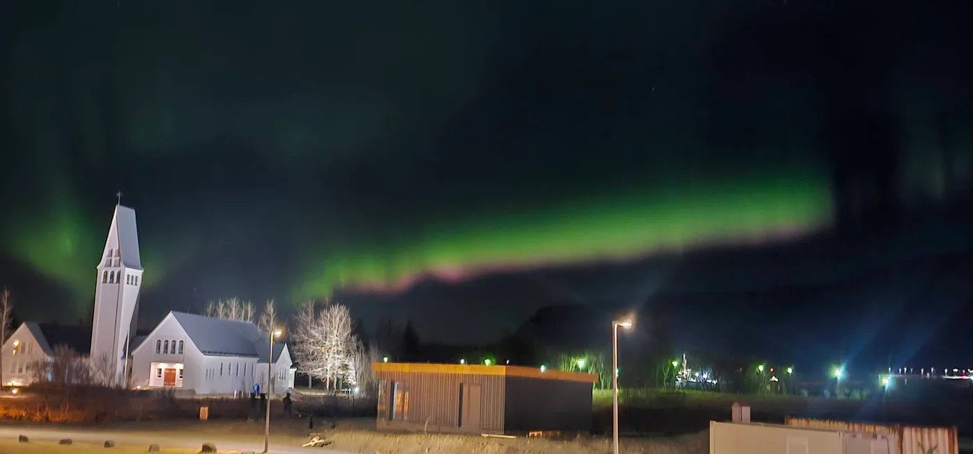 A green aurora band over a white church with a tower, lit by streetlights