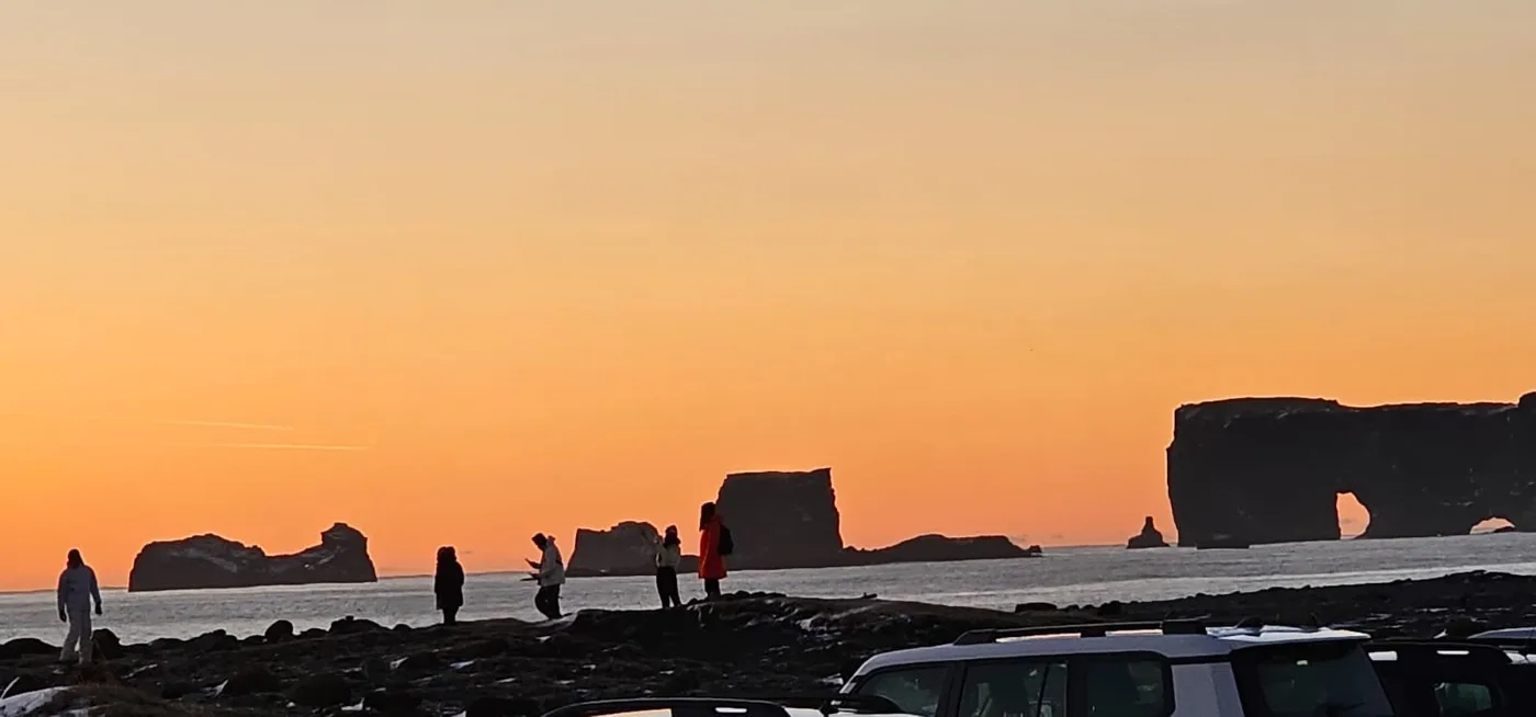 Silhouettes of people on a black beach at sunset, the Dyrhólaey rock arch behind them and a car to the right