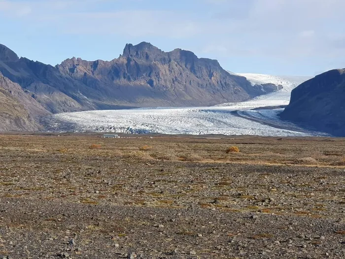 A glacier tongue spilling down between dark mountains onto a flat gravel plain