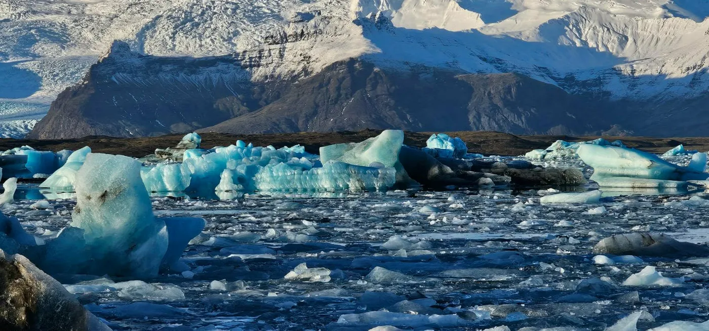 Broken ice filling the lagoon under a snow-streaked mountain and blue sky