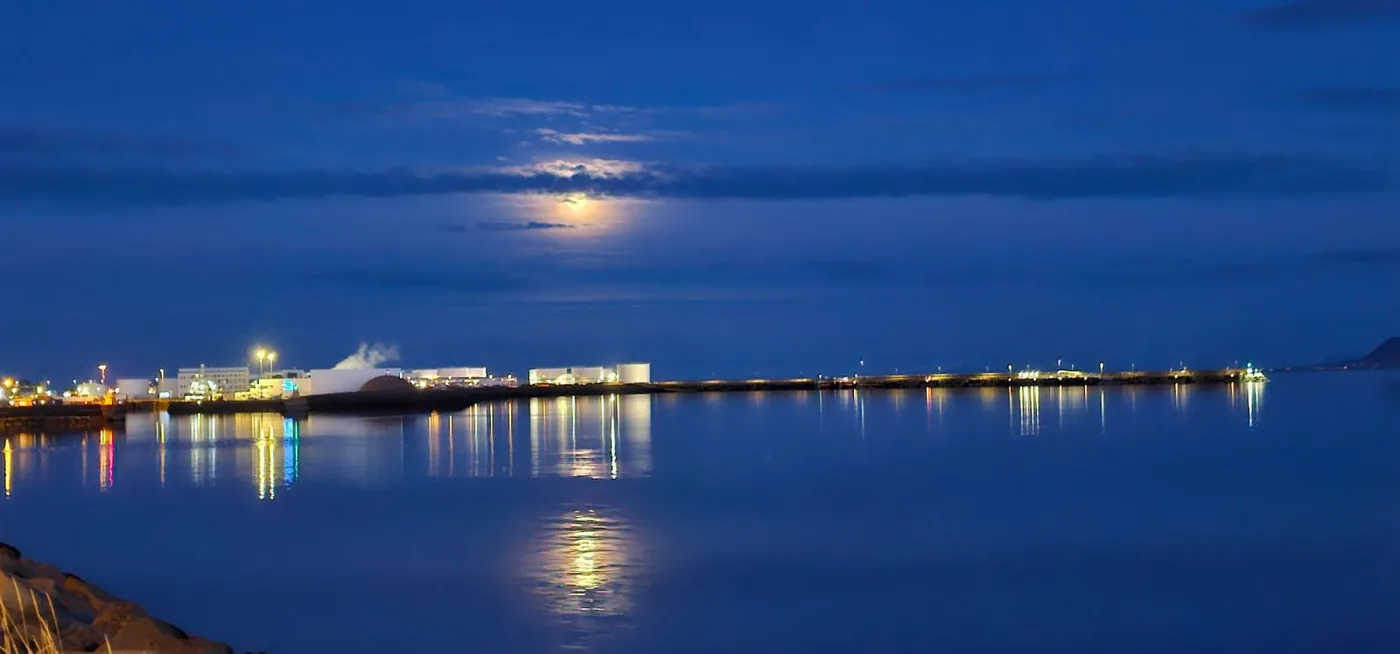 A full moon in a deep blue sky over a bay, town lights strung along the far shore and reflected in the water