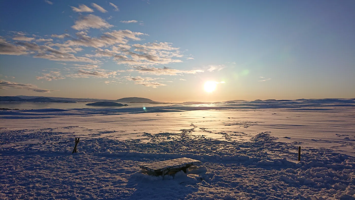 The winter sun low over a frozen lake, a bench in the snow in the foreground